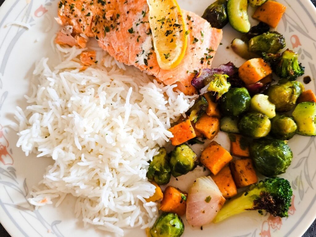 Healthy dinner plate with baked salmon topped with lemon, fluffy white rice, and roasted vegetables including Brussels sprouts, zucchini, sweet potato, and broccoli.