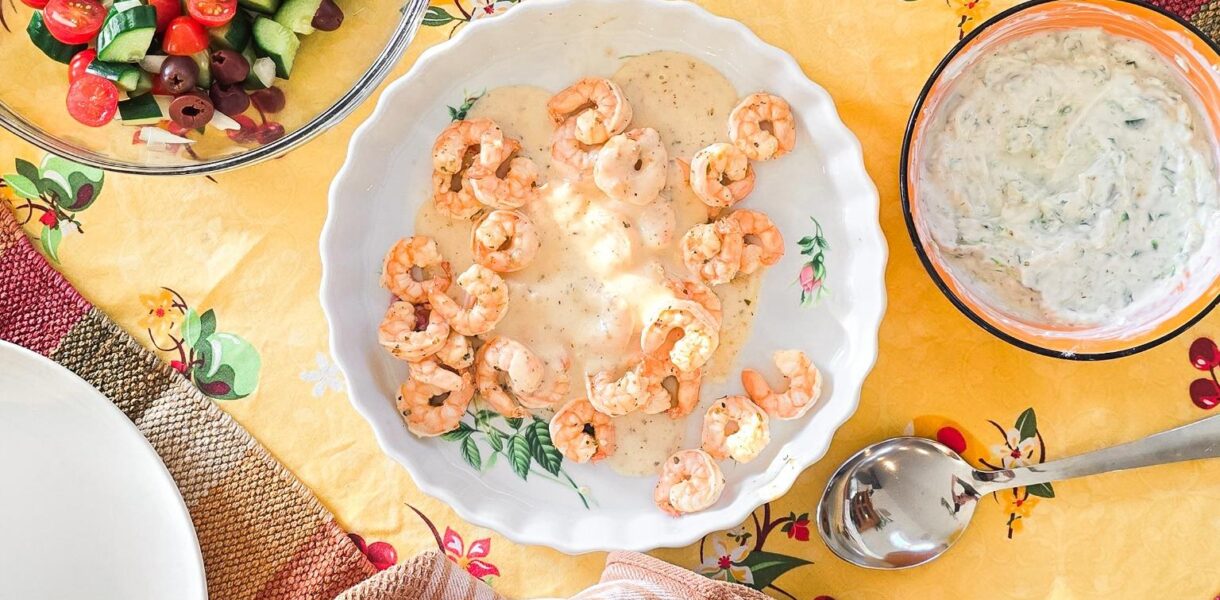 A Mediterranean-style dinner spread featuring grilled shrimp in a creamy lemon sauce, fresh Greek salad with cucumbers, tomatoes, and olives, diced red bell peppers, tzatziki dip, and warm pita bread, all served on a vibrant yellow floral tablecloth.