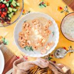 A Mediterranean-style dinner spread featuring grilled shrimp in a creamy lemon sauce, fresh Greek salad with cucumbers, tomatoes, and olives, diced red bell peppers, tzatziki dip, and warm pita bread, all served on a vibrant yellow floral tablecloth.