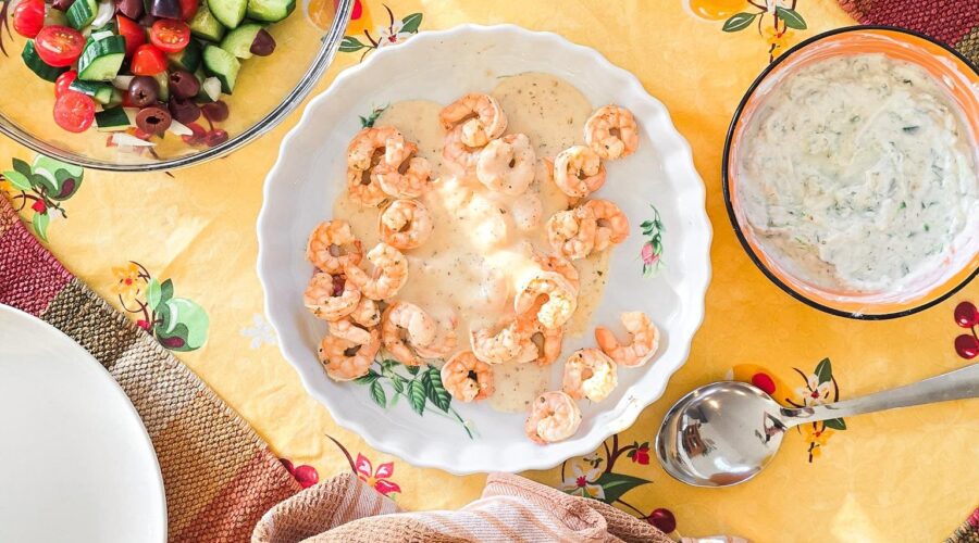 A Mediterranean-style dinner spread featuring grilled shrimp in a creamy lemon sauce, fresh Greek salad with cucumbers, tomatoes, and olives, diced red bell peppers, tzatziki dip, and warm pita bread, all served on a vibrant yellow floral tablecloth.