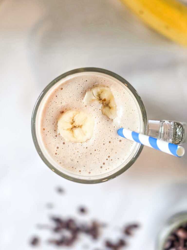 Top-down view of a coffee banana protein smoothie in a glass mug, garnished with banana slices and a striped straw, with scattered coffee beans and a banana in the background.