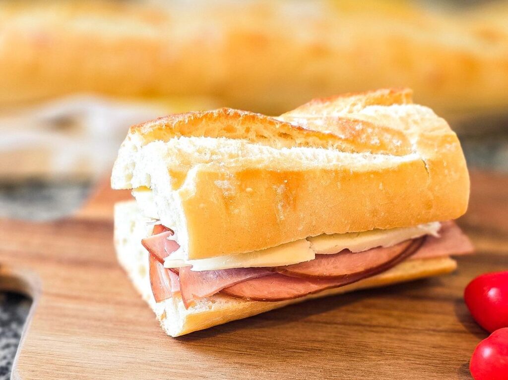 French-style ham and cheese sandwich on a crusty baguette, served on a wooden board with cherry tomatoes.