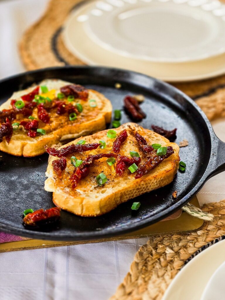 Angled view of two slices of egg-in-a-hole sourdough toast in a cast iron skillet, topped with sundried tomatoes and chopped green onions. A set table with cream-colored plates and woven placemats provides a cozy Mediterranean-inspired backdrop.