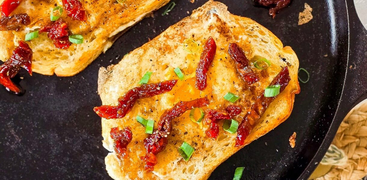 Overhead view of two slices of sourdough toast with sunny-side-up eggs cooked in the center, garnished with sundried tomatoes and chopped green onions, served in a black cast iron skillet on a woven placemat.