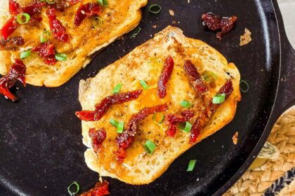 Overhead view of two slices of sourdough toast with sunny-side-up eggs cooked in the center, garnished with sundried tomatoes and chopped green onions, served in a black cast iron skillet on a woven placemat.