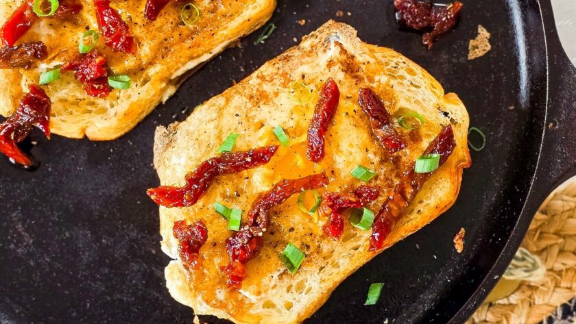 Overhead view of two slices of sourdough toast with sunny-side-up eggs cooked in the center, garnished with sundried tomatoes and chopped green onions, served in a black cast iron skillet on a woven placemat.