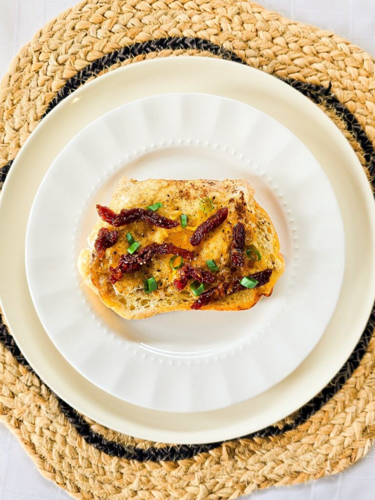 Overhead view of a single slice of sourdough egg-in-a-hole toast topped with sundried tomatoes and chopped green onion, served on a layered white plate over a woven placemat.