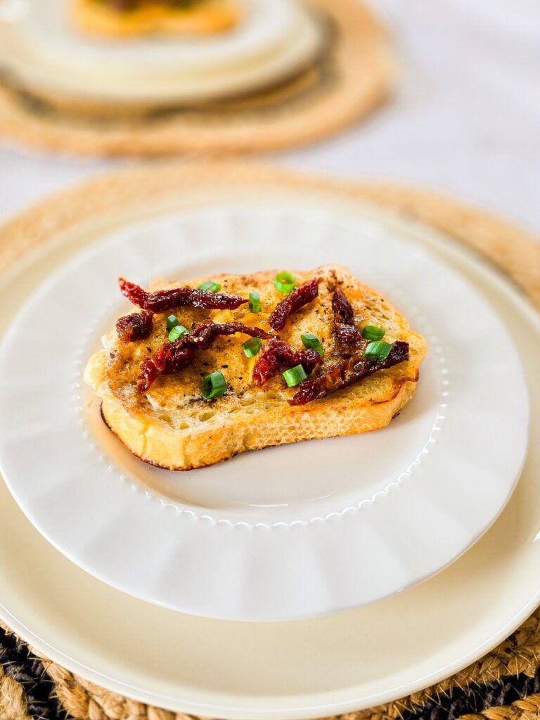 Close-up of a single slice of egg-in-a-hole toast garnished with sundried tomatoes and green onions, served on a white plate with a woven charger underneath. Another plate with toast is softly blurred in the background, creating a warm, inviting breakfast scene.