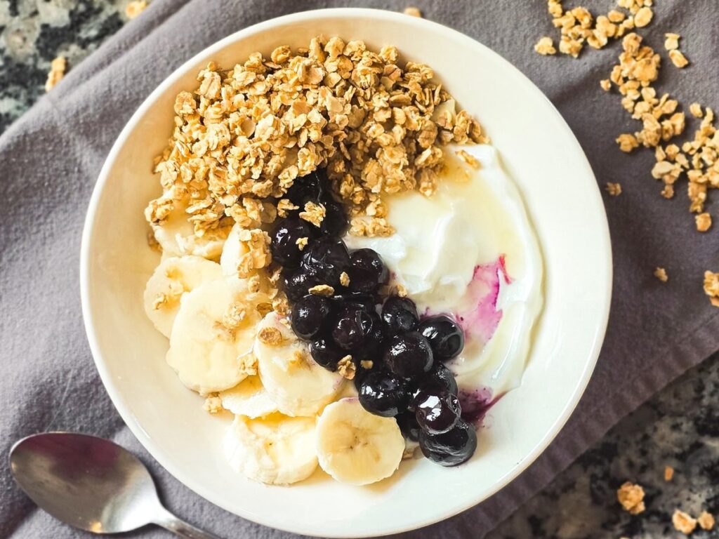 High-protein yogurt bowl topped with banana slices, blueberry compote, and crunchy granola on a grey napkin with a spoon.
