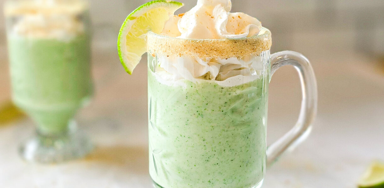 Two glass mugs filled with a creamy green key lime smoothie, topped with whipped cream and garnished with lime wedges and a graham cracker rim, sitting on a white tablecloth.