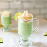 Two glass mugs filled with a creamy green key lime smoothie, topped with whipped cream and garnished with lime wedges and a graham cracker rim, sitting on a white tablecloth.