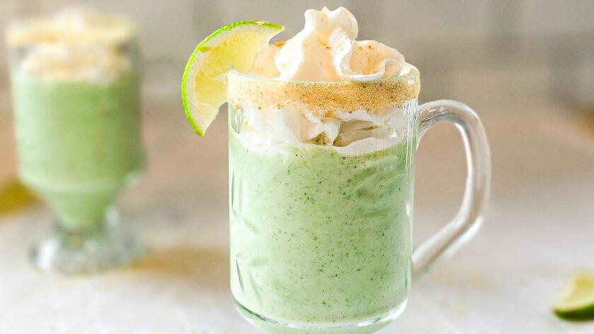 Two glass mugs filled with a creamy green key lime smoothie, topped with whipped cream and garnished with lime wedges and a graham cracker rim, sitting on a white tablecloth.