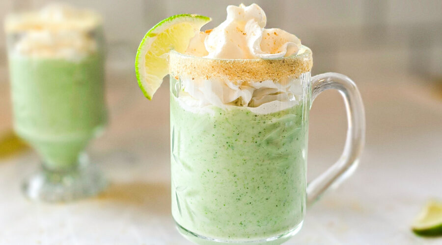Two glass mugs filled with a creamy green key lime smoothie, topped with whipped cream and garnished with lime wedges and a graham cracker rim, sitting on a white tablecloth.