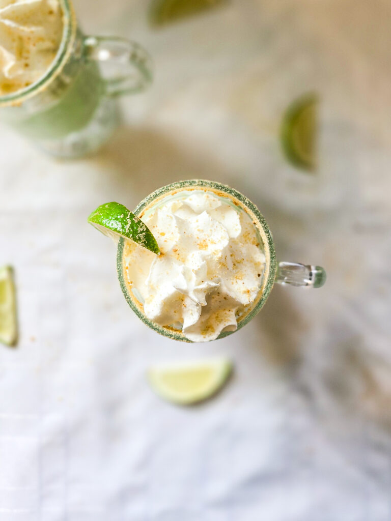 Overhead view of a whipped cream-topped smoothie with a graham cracker rim and lime wedge, styled on a white tablecloth with scattered lime slices for a fresh summer vibe.
