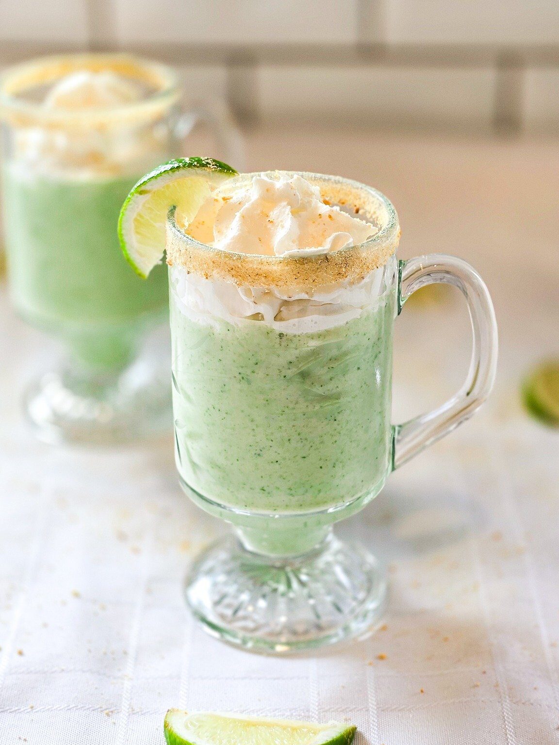 A vibrant green key lime smoothie served in a glass mug, topped with whipped cream, a lime wedge, and a graham cracker rim—perfect for summer mornings or a healthy dessert alternative.