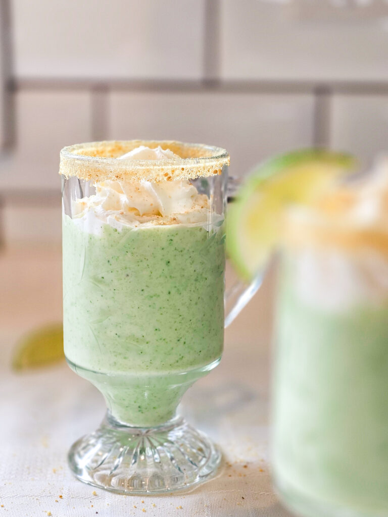 Creamy key lime smoothie with flecks of green, served in a pedestal glass and garnished with whipped cream and a graham cracker crusted rim, captured in soft natural light.