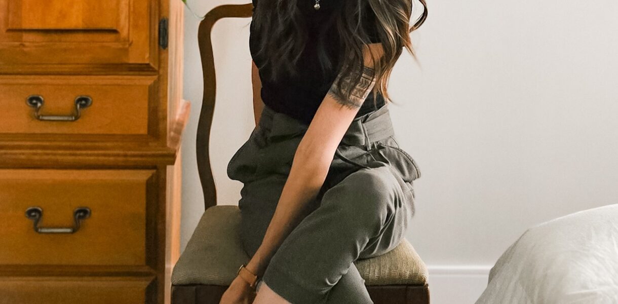 Woman sitting on a wooden chair in a neutral-toned bedroom, wearing a black top, high-waisted olive trousers, and black heels, reaching to adjust her shoe.