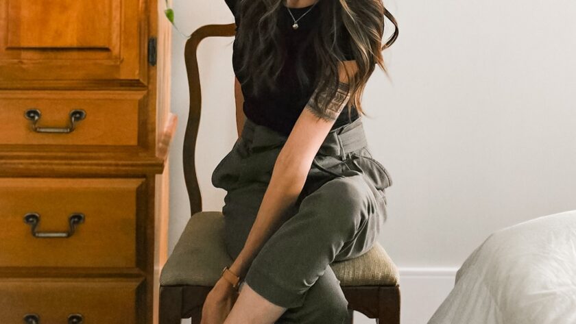 Woman sitting on a wooden chair in a neutral-toned bedroom, wearing a black top, high-waisted olive trousers, and black heels, reaching to adjust her shoe.