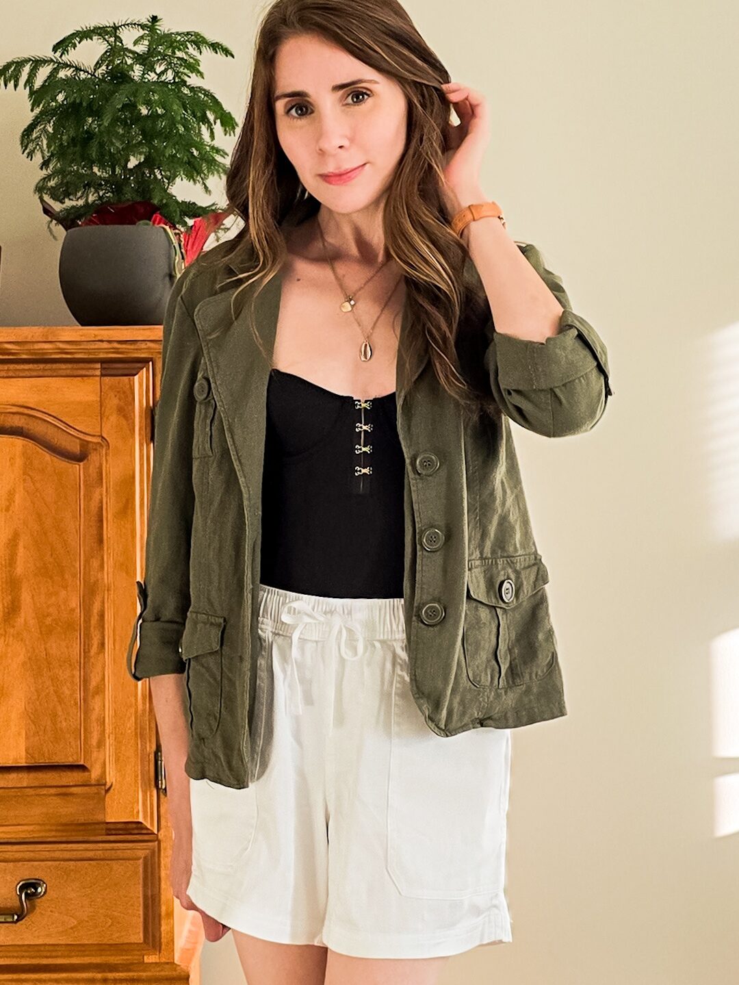 Woman standing indoors wearing a dark green utility jacket over a black top and high-waisted white drawstring shorts, with soft natural light illuminating the space.