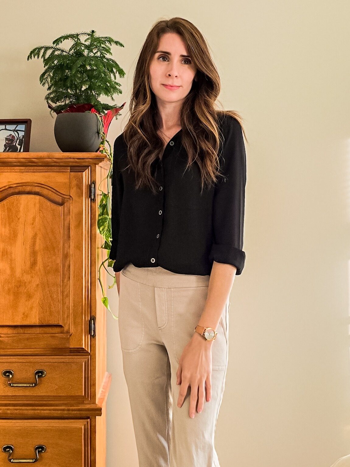 Woman standing in a softly lit room wearing a casual black button-up shirt and beige pants, showcasing a neutral-toned, minimalist outfit.