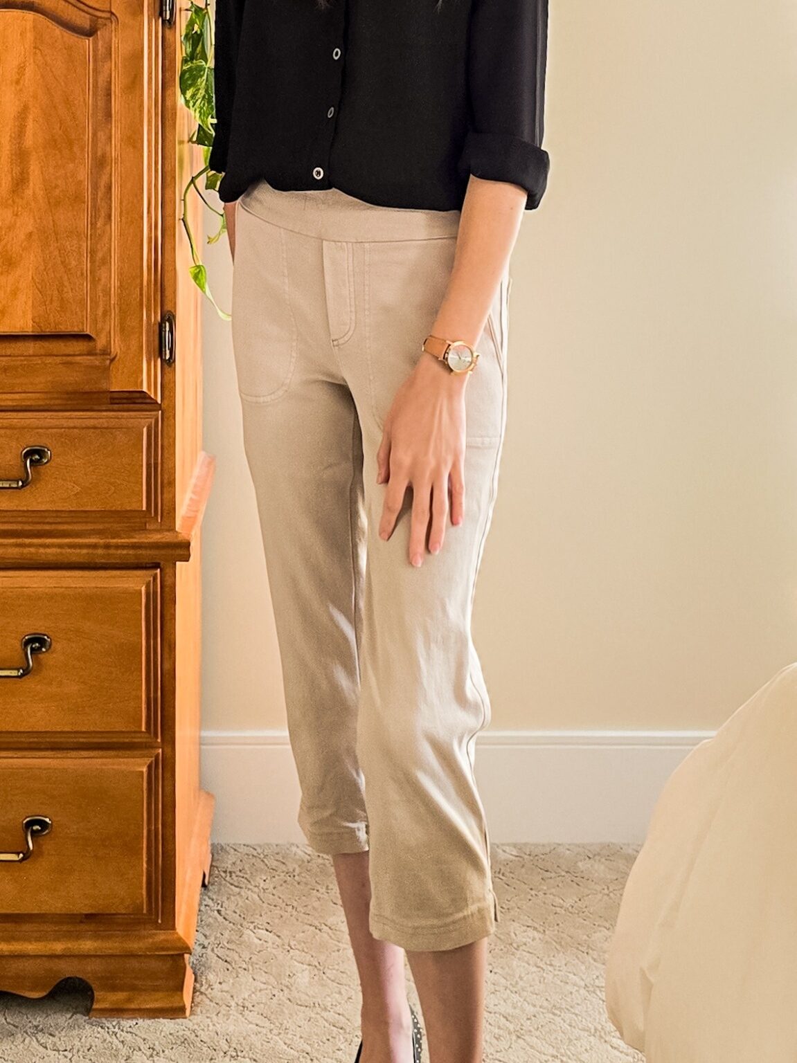 Woman standing beside a wooden dresser, wearing a black blouse with rolled sleeves and beige cropped pants, styled with a tan watch and black heels.