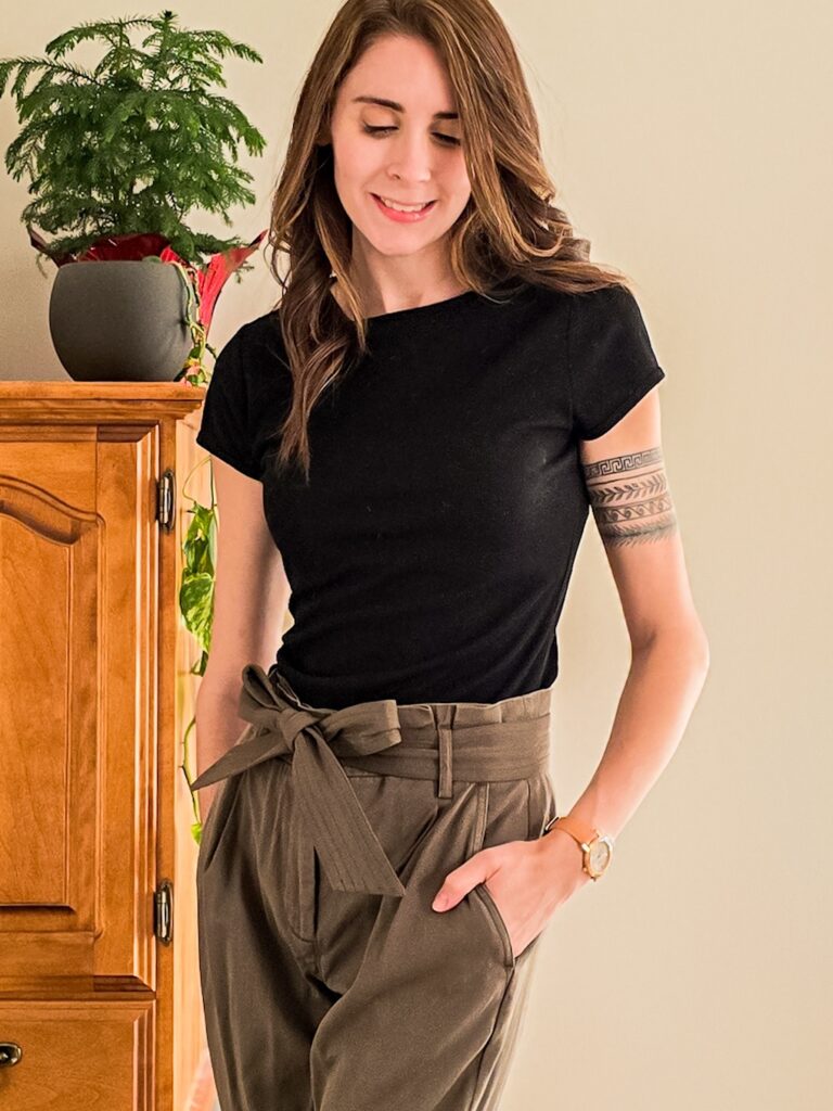 Woman wearing an all-neutral outfit featuring a black fitted tee and high-waisted olive paperbag pants, accessorized with a tan watch and subtle gold jewelry.